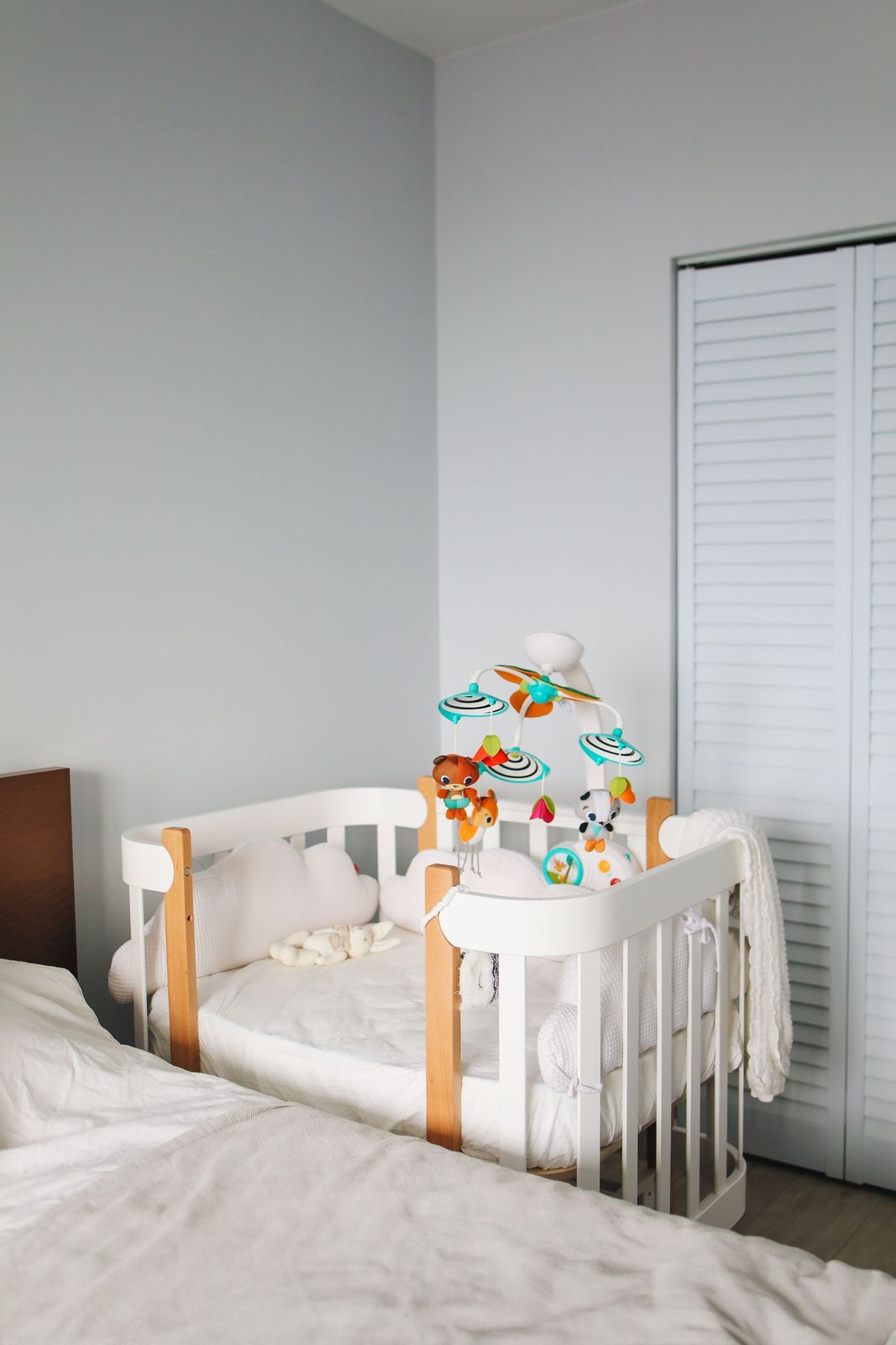 White bedside co-sleeper crib with natural wood posts and a colourful mobile beside a parent bed