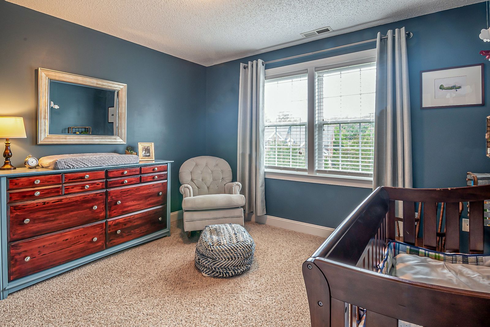 Blue nursery with a red wood dresser used as a changing station, a tufted glider chair and a dark wood crib