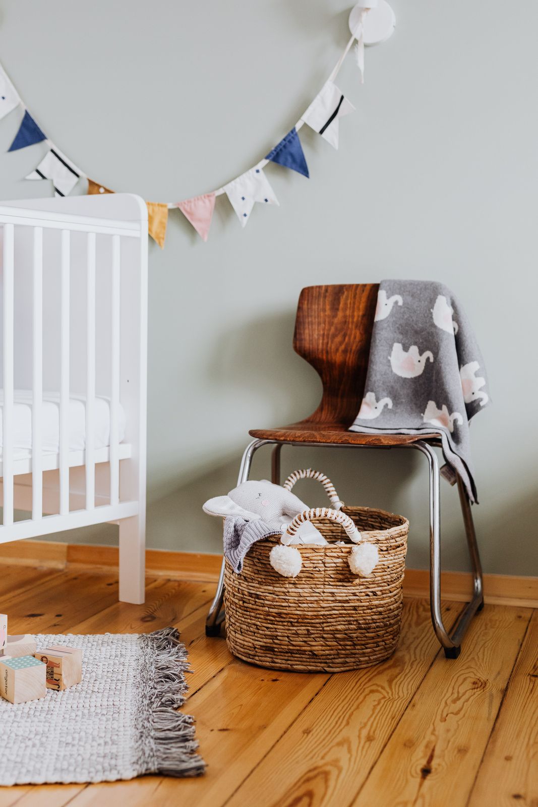Nursery corner with a white crib, wooden chair with elephant blanket, woven basket and fabric bunting