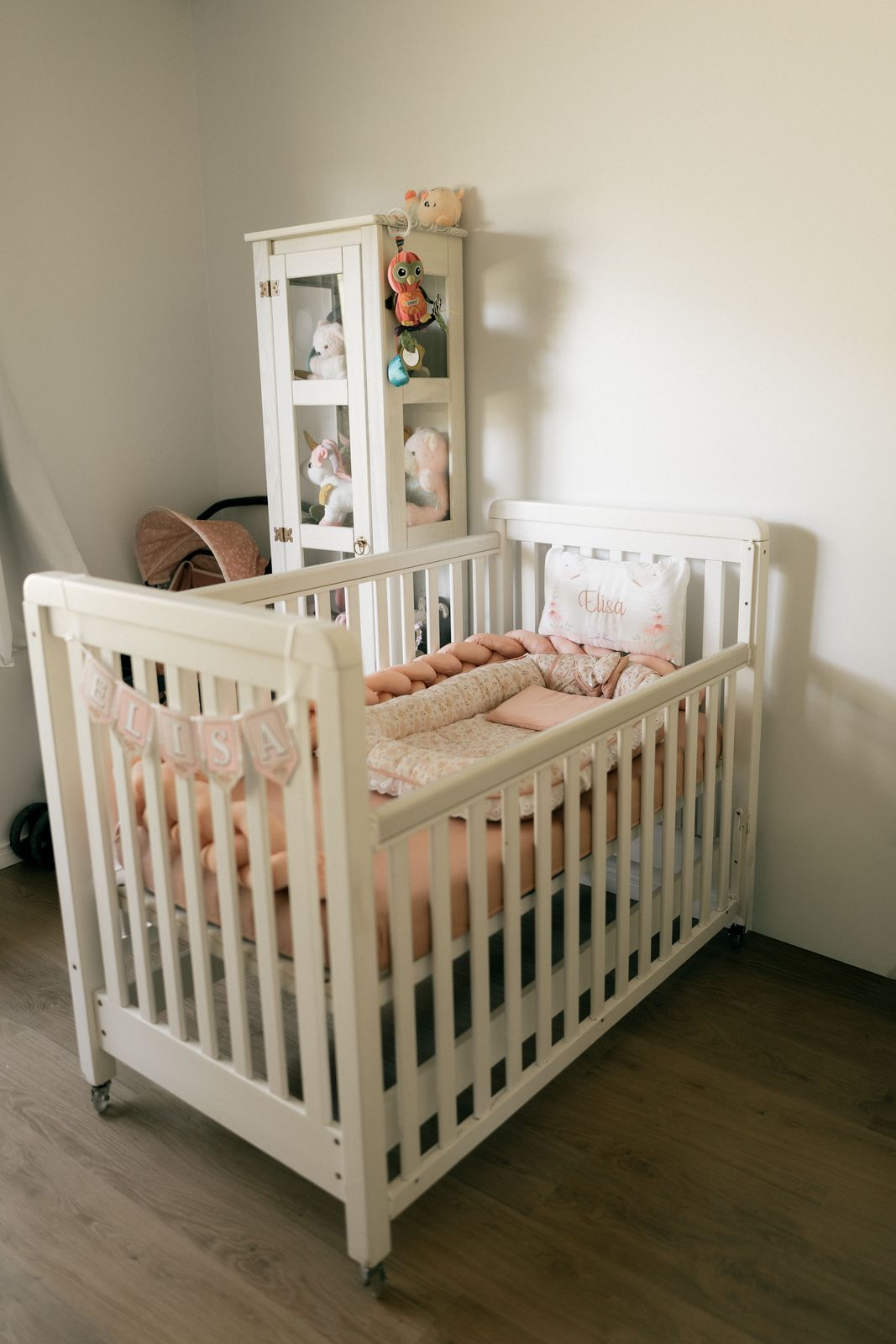 White crib on castors with peach bedding, a name banner and a tall white display cabinet behind